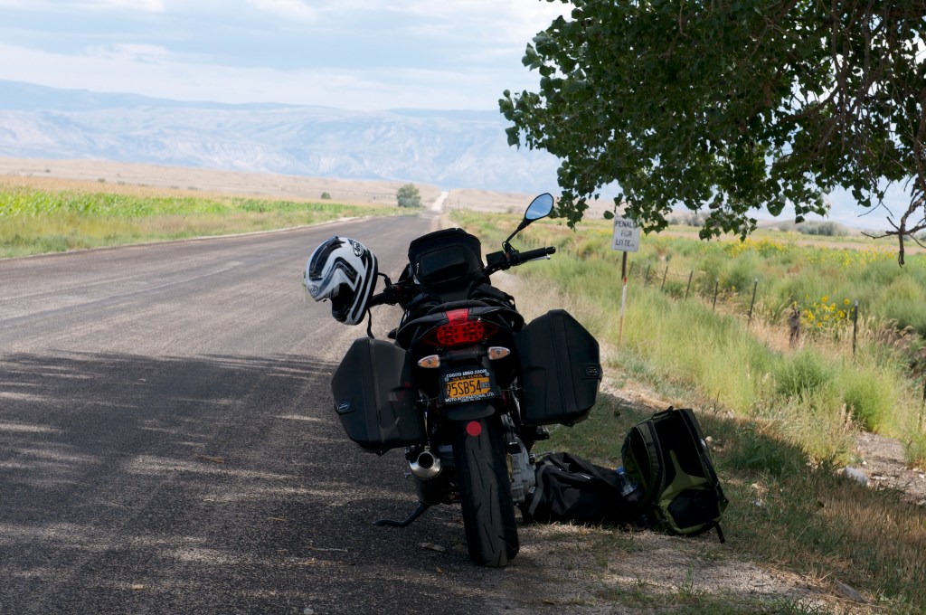 A parked motorcycle with a helmet and a bag beside it on a rural road with distant mountains in the background.